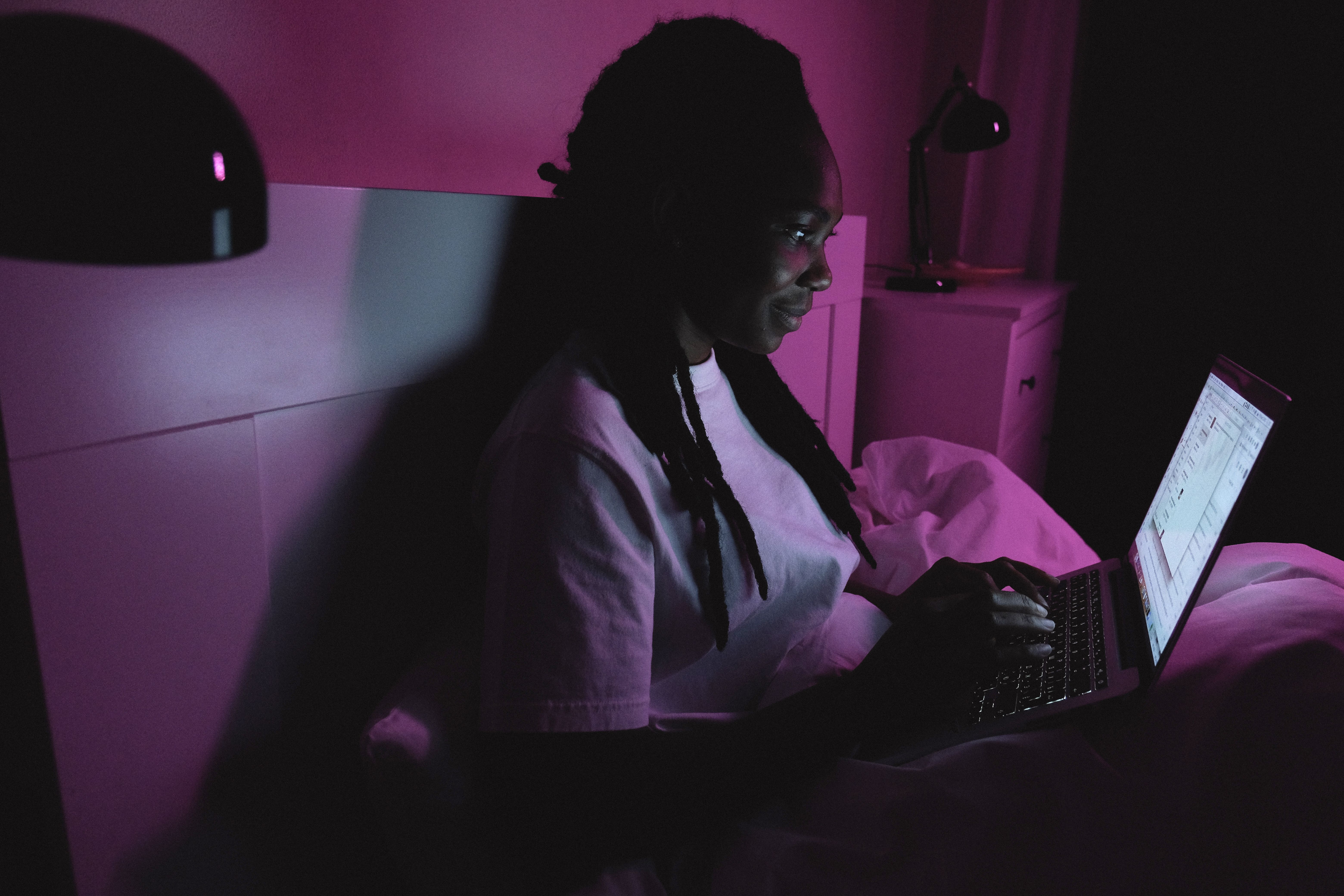 Woman using a laptop in bed under purple lighting, evoking a private late-night digital connection.